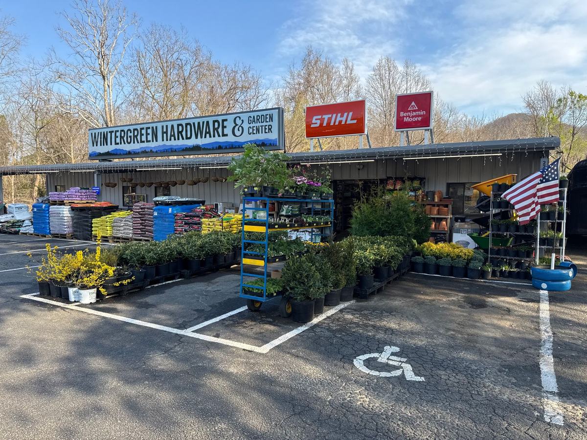 The storefront on a clear spring day: shrubs and flowering plants set out on the lot, bagged soil and mulch stacked at left, and the store sign with Stihl and Benjamin Moore signs on the roof